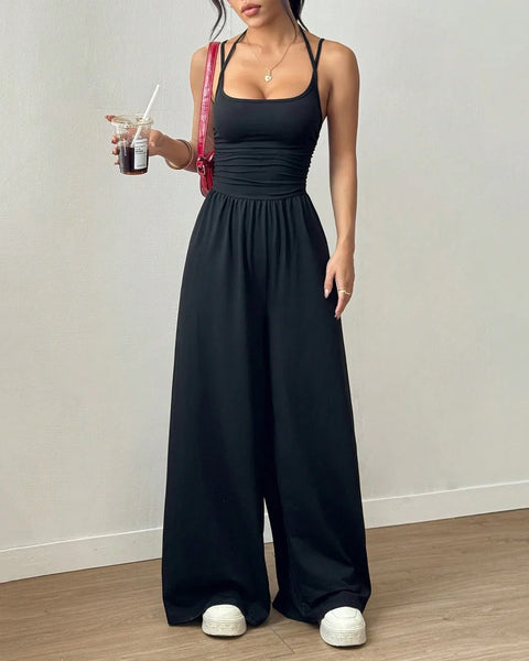 A woman in a black sleeveless wide-leg jumpsuit holds a drink, standing confidently with a casual yet stylish demeanor.