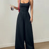 A woman in a black sleeveless wide-leg jumpsuit holds a drink, standing confidently with a casual yet stylish demeanor.

