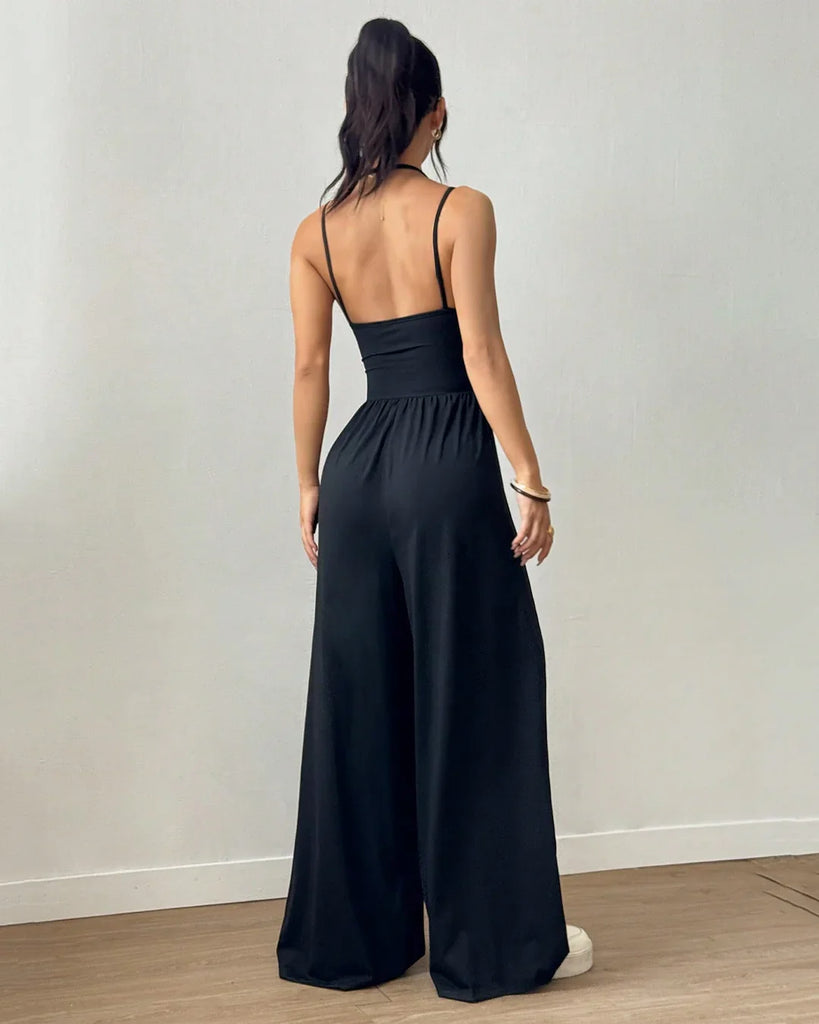 A woman in a black sleeveless wide-leg jumpsuit holds a drink, standing confidently with a casual yet stylish demeanor.