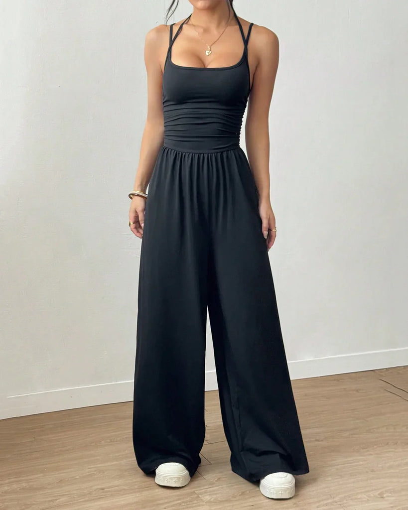 A woman in a black sleeveless wide-leg jumpsuit holds a drink, standing confidently with a casual yet stylish demeanor.