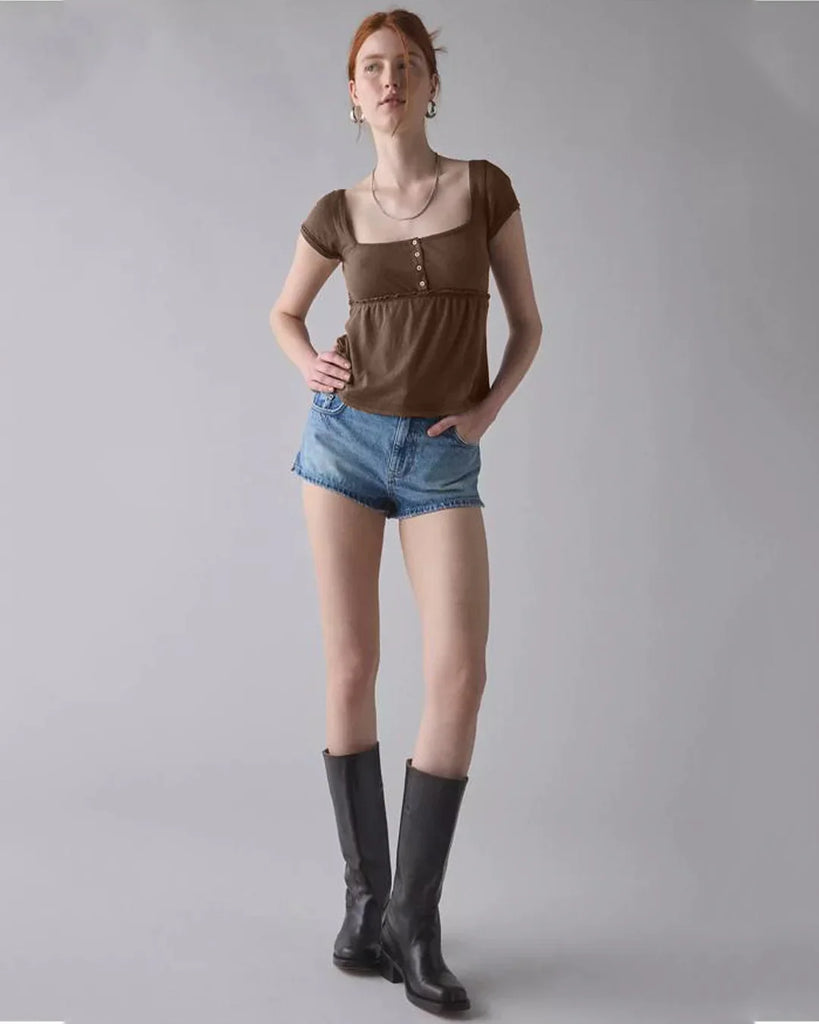  A woman in a brown button-front square neck top and shorts, standing confidently with a neutral background.
