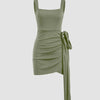 Olive green mini dress with ruched fabric, featuring a knotted tie, square neck, and sleeveless design.
