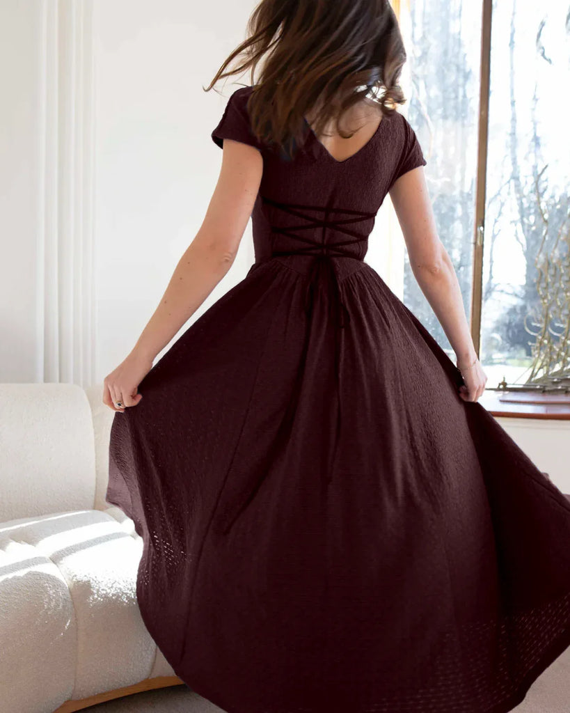 Back view of a woman poses in a maroon cap sleeve fit and flare midi dress, showcasing its elegant silhouette against a neutral background.