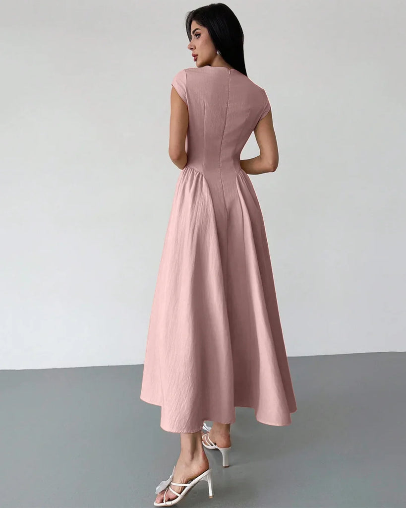 Back view of a  woman in a powder pink one-shoulder flared hem midi dress takes a selfie, smiling at the camera.