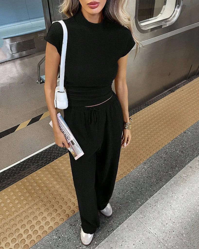 A woman in a black short sleeve top and wide-leg trousers stands confidently on a train platform.