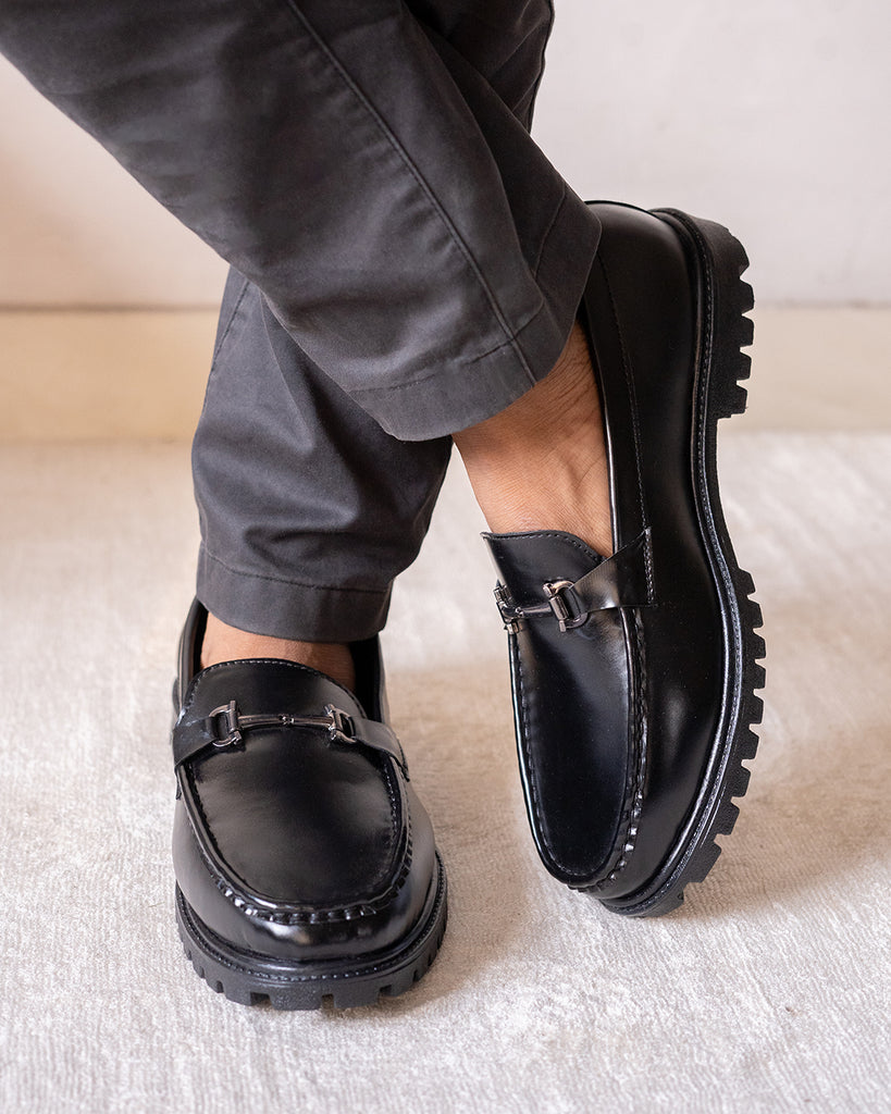 A model standing with cross leg wearing black slip on Chunky loafers with buckle detail