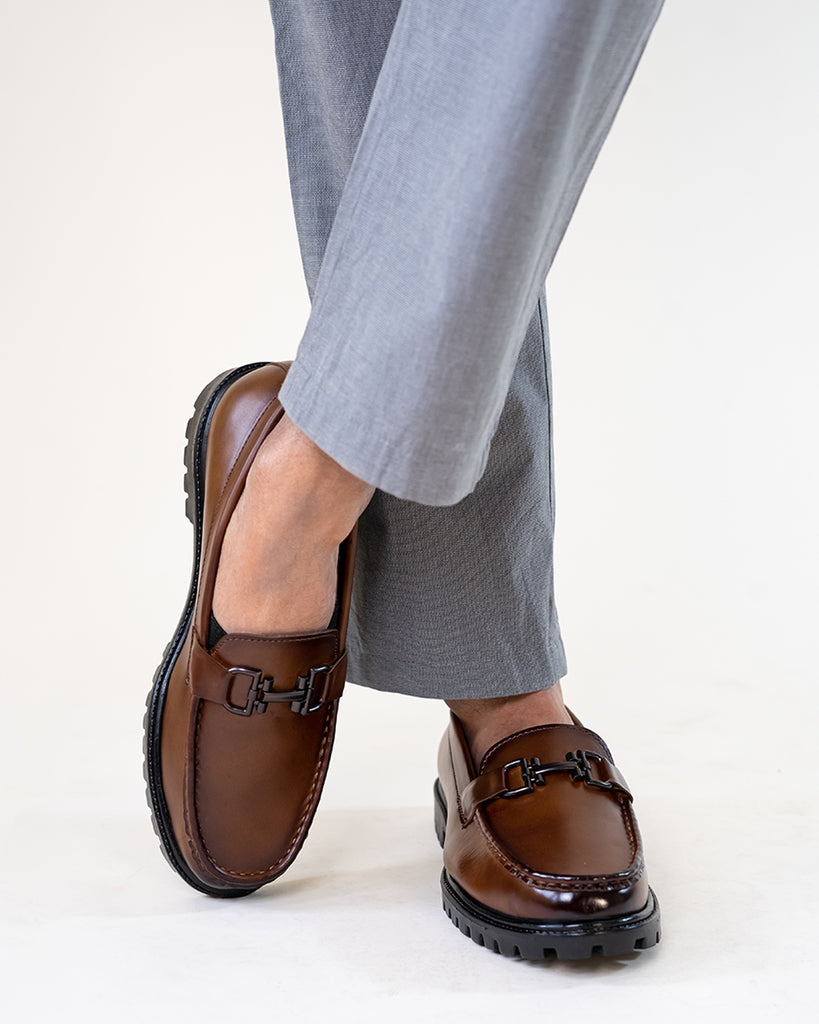 A model crossing his leg wearing grey pants and chunky brown loafers 